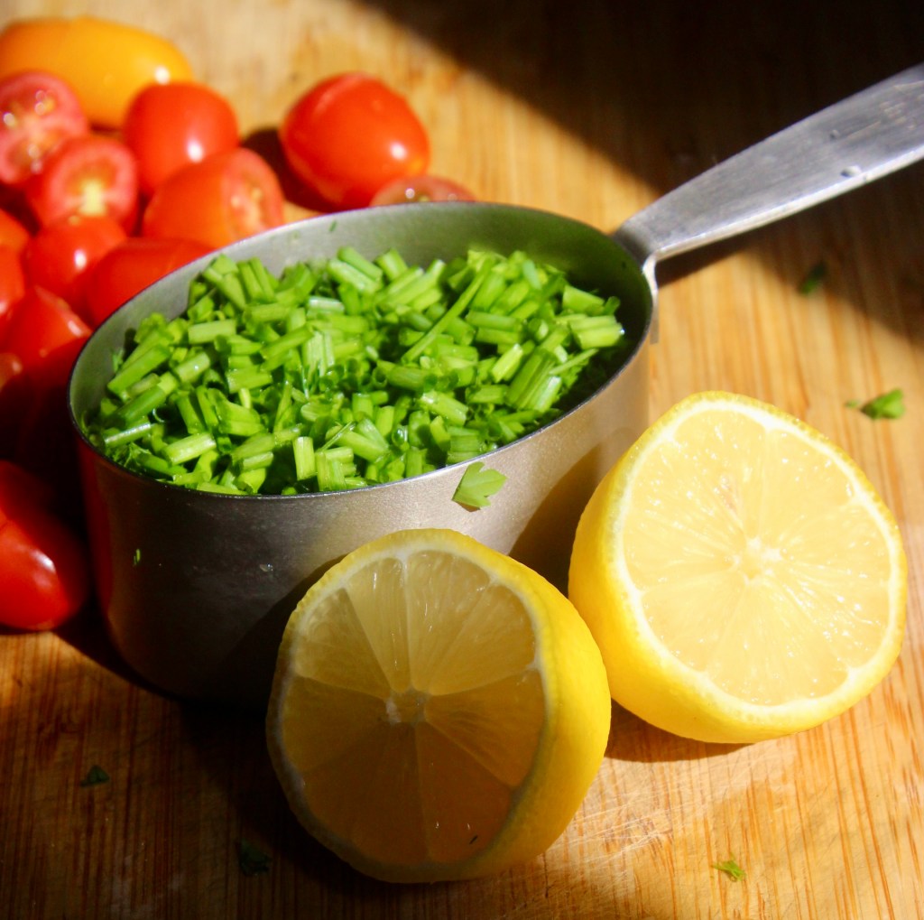 Chopped green herbs in a metal measuring cup, surrounded by halved lemons and cherry tomatoes on a wooden cutting board.