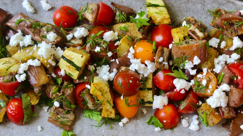 A colorful platter of roasted vegetables including cherry tomatoes, zucchini, eggplant, and herbs, topped with crumbled feta cheese.