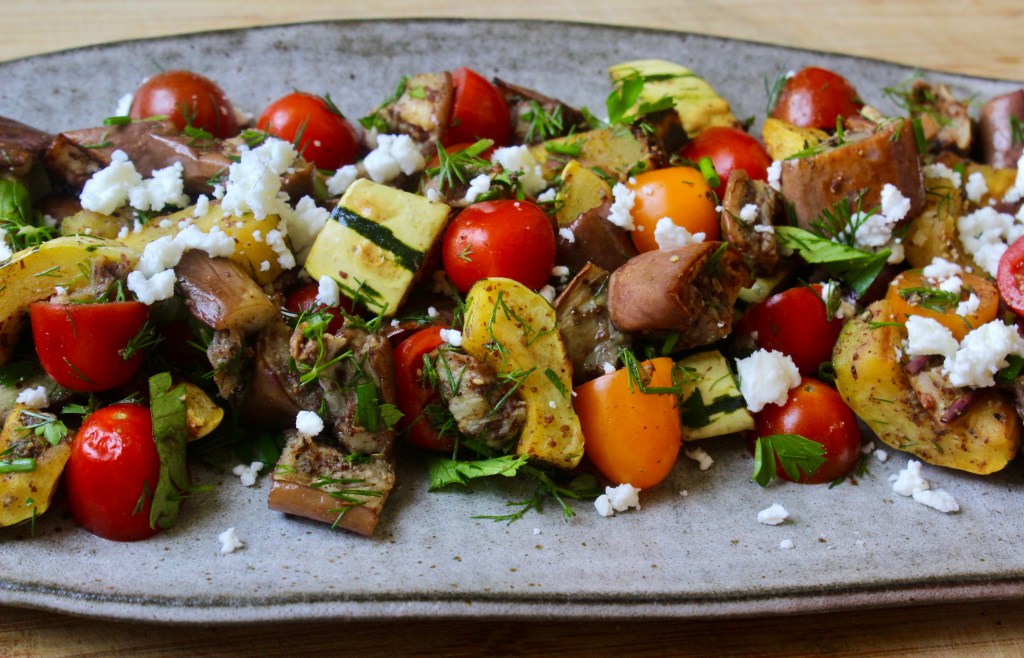 A colorful platter of roasted vegetables including potatoes, cherry tomatoes, and zucchini, topped with crumbled feta and fresh herbs.